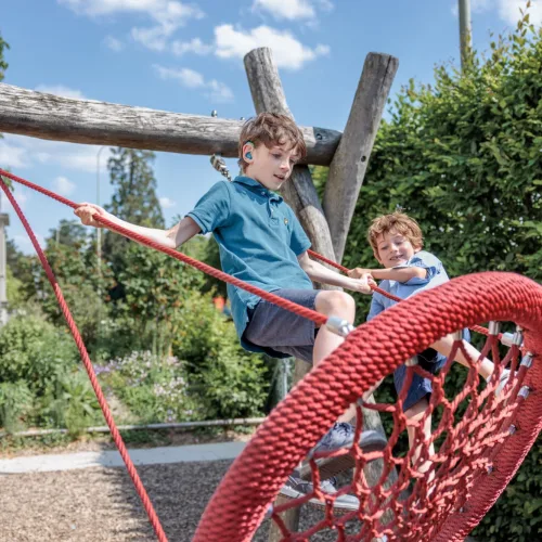 Zwei Jungen klettern auf einem Spielplatz über ein rotes Kletternetz, der ältere trägt ein blaues Hörgerät hinter dem Ohr.